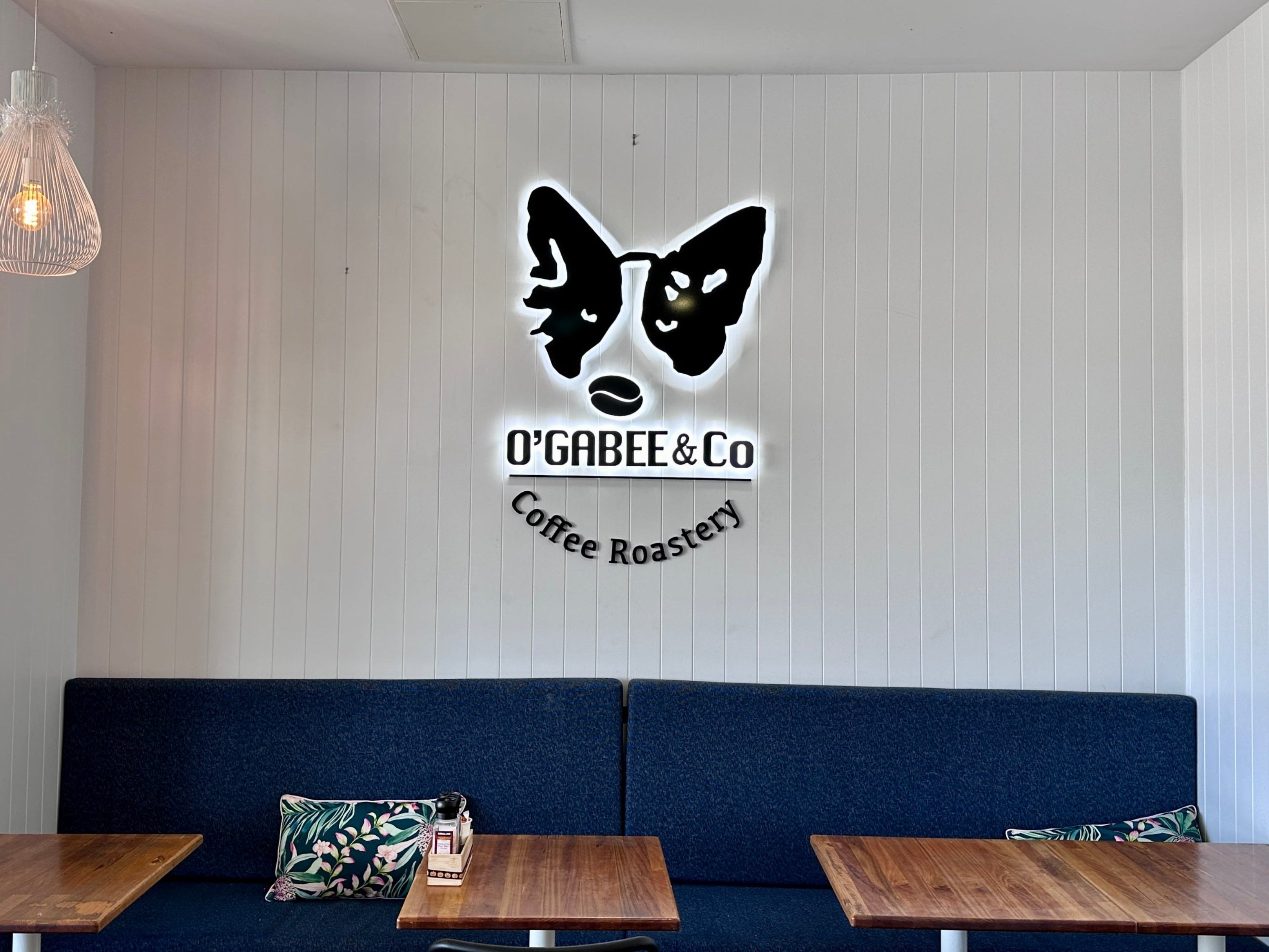 A restaurant or coffee shop interior with a long navy blue booth against a white paneled wall. The wall features a black butterfly logo with the text 'O'GABEE & Co Coffee Roastery'.