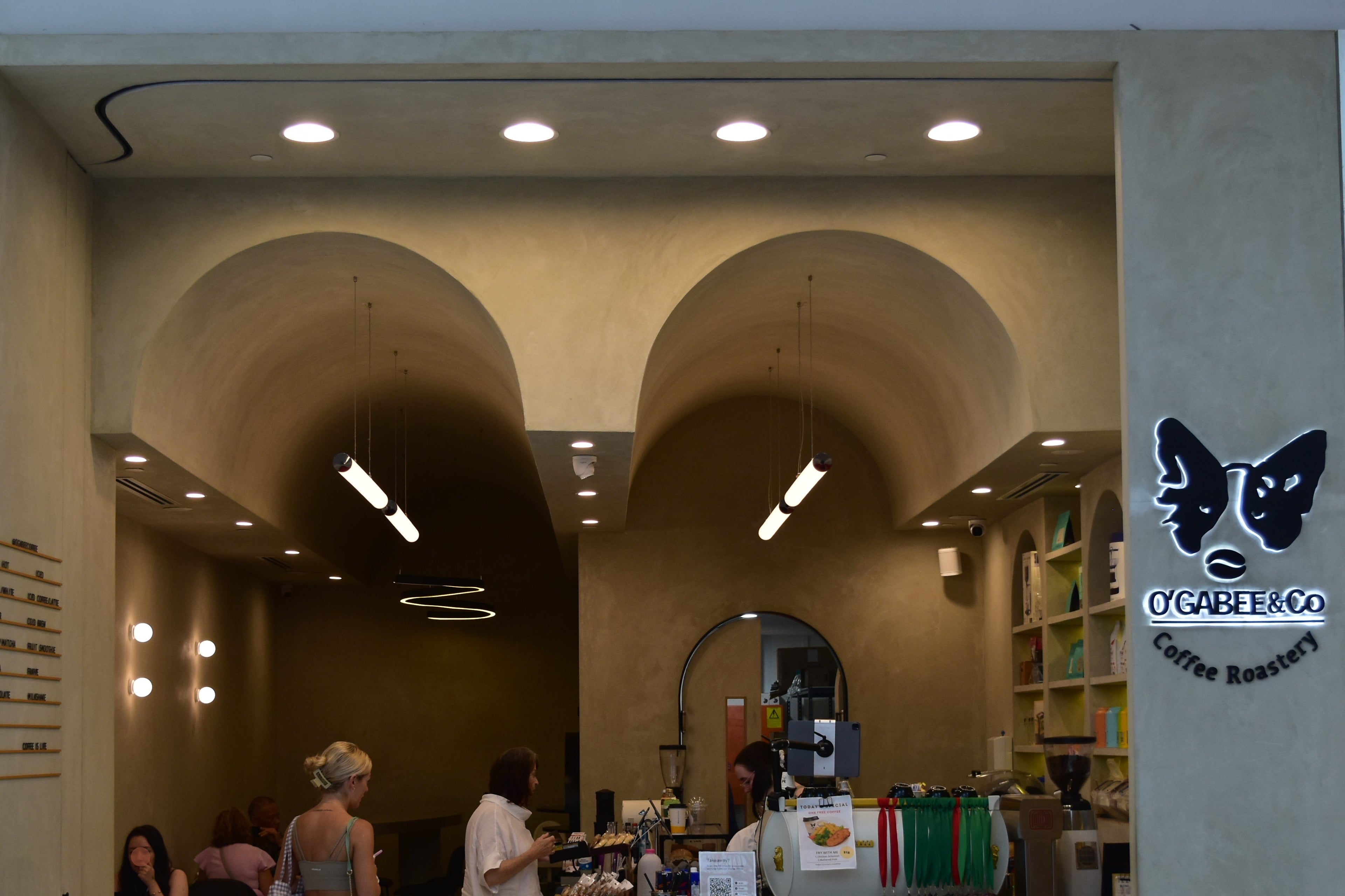 A storefront view of a cafe, coffee shop with arched interior walls, a tiled counter, and several people standing inside at Chermsdie Westfield.