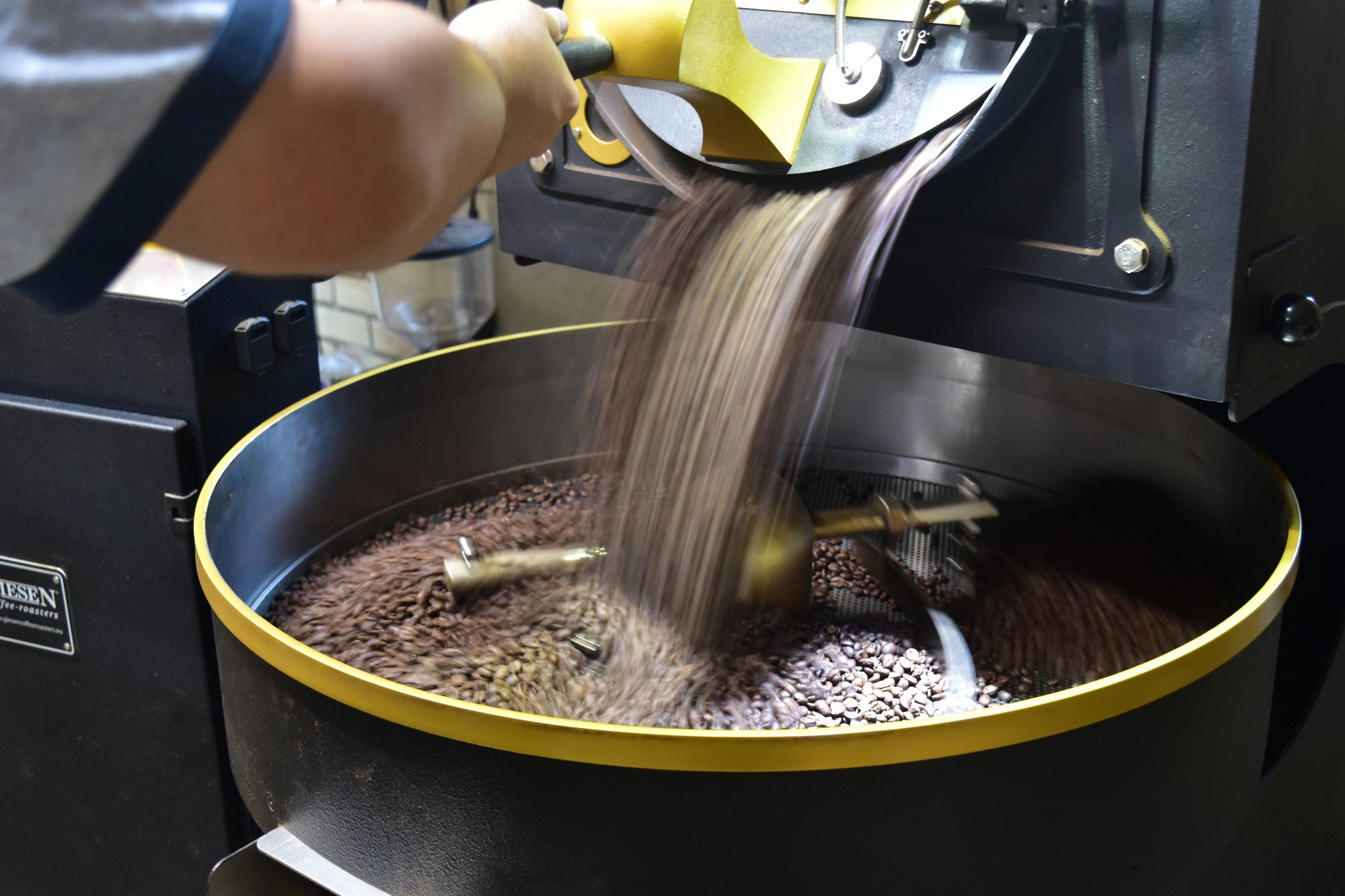 Coffee beans being poured from a metal hopper into a black coffee roaster bowl with a gold rim, with a person's arm visible operating the machine.