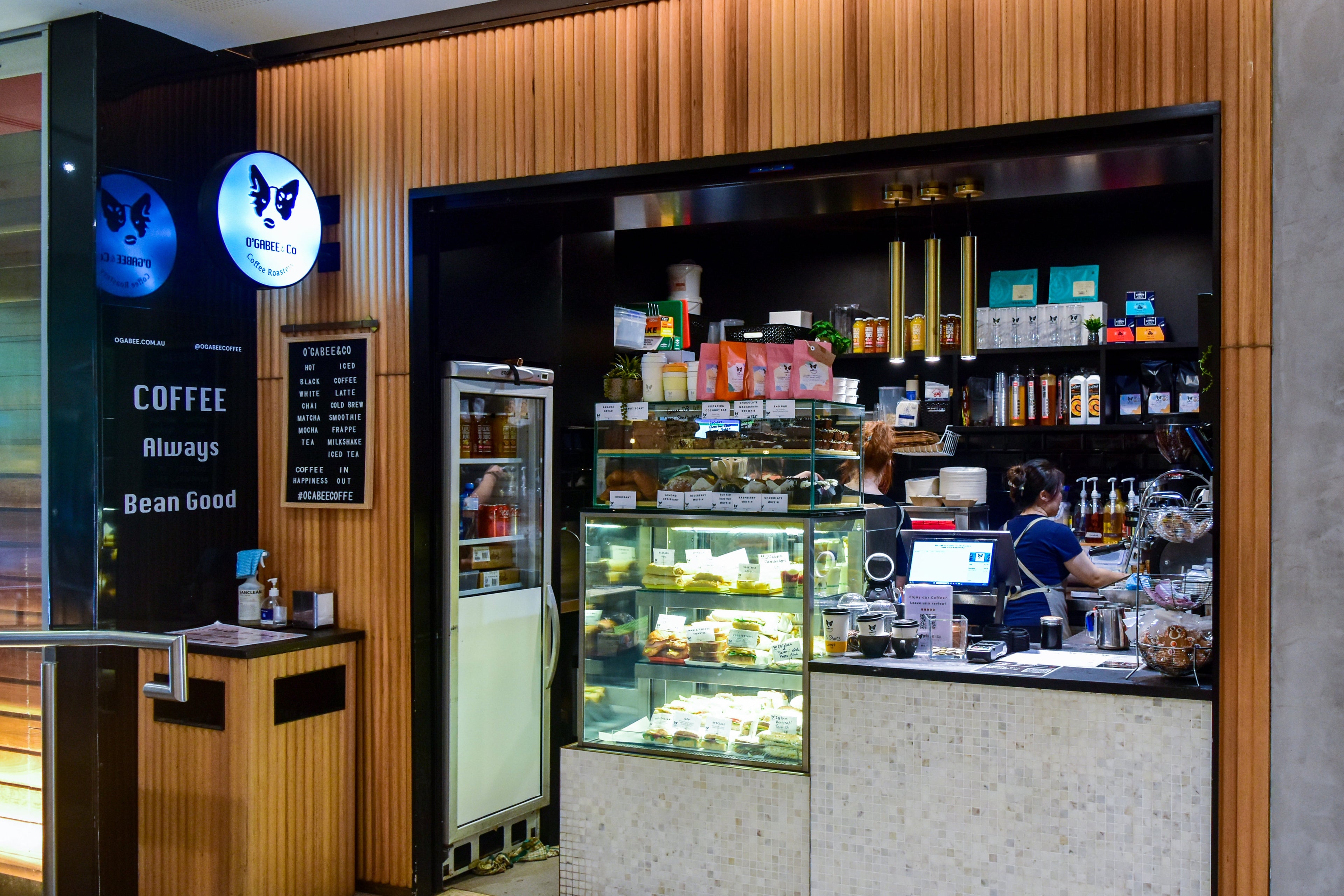 A coffee shop storefront with wood-paneled walls, a glass display case filled with pastries, a counter, and a staff member working inside. Signs on the left read 'COFFEE Always Bean Good' and show butterfly logos.